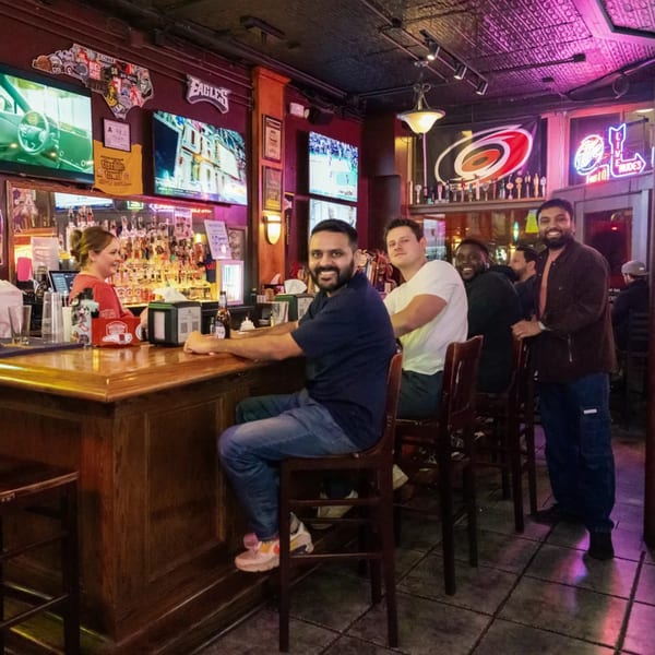 Patrons and a bartender at the Woody's @ City Market bar, with multiple sports screens and neon signs.