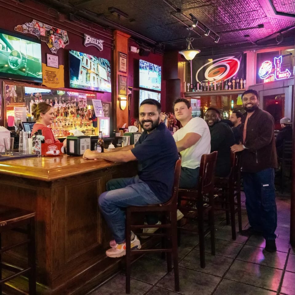 Patrons and a bartender at the Woody's @ City Market bar, with multiple sports screens and neon signs.
