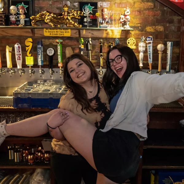 Two smiling women pose playfully in front of beer taps at Woody's @ City Market sports bar.