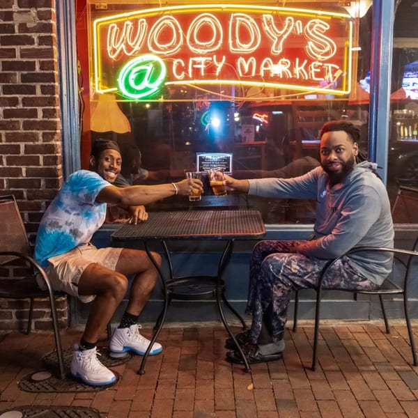 Two men toast beers at an outdoor table in front of Woody's @ City Market neon sign.