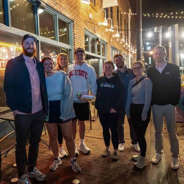 A group of eight young adults smiling outside Woody's @ City Market at night.