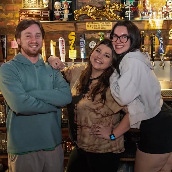 Three smiling people pose in front of beer taps and sports memorabilia at Woody's @ City Market.