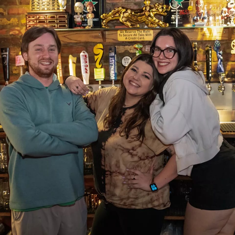 Three smiling people pose in front of beer taps and sports memorabilia at Woody's @ City Market.