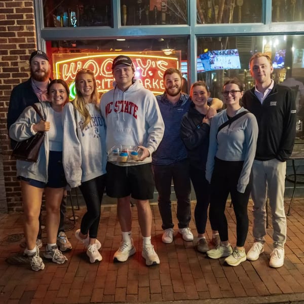 Group of eight young adults smiling outside Woody's @ City Market, a sports bar with neon signs.