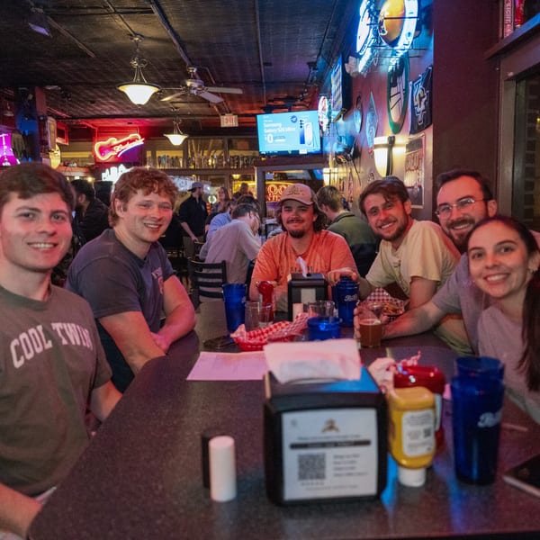 Six young adults smiling at a table in a dimly lit sports bar with neon signs.