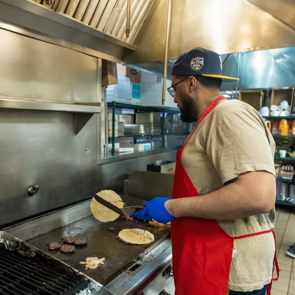 A cook in a red apron and baseball cap prepares food on a flat-top grill in a commercial kitchen.