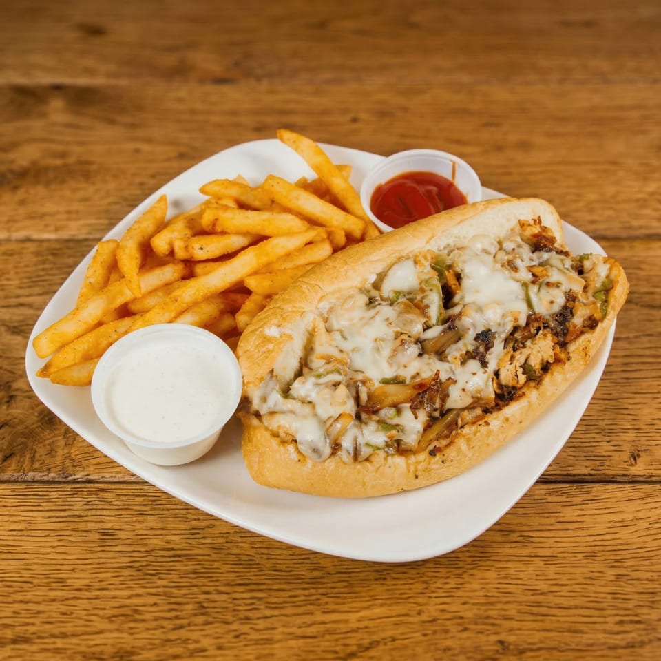 Cheesesteak sandwich with melted cheese, onions, and peppers, served with fries, ranch, and ketchup.